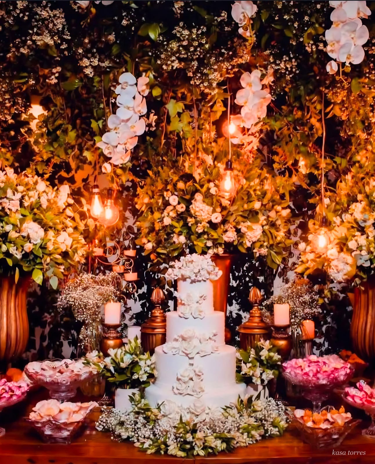 Mesa de bolo de casamento branco entre folhagens, orquídeas e luminárias edison pendentes — Kasa Torres